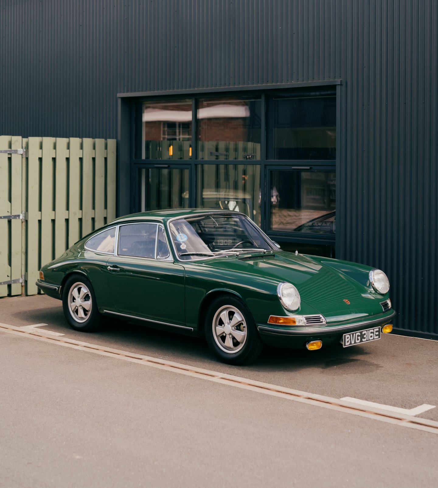 A side on view of a dark green classic porsche in front of a dark grey building, making the car stand out