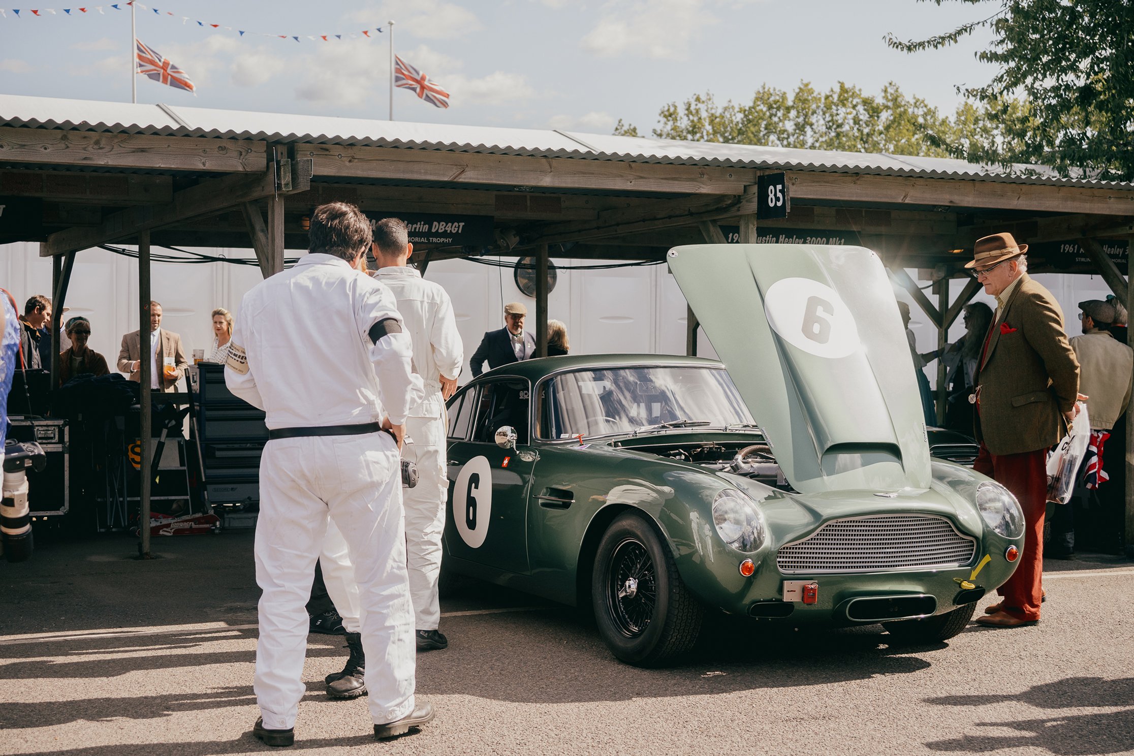 Thee men stand looking at an green aston martin racing car, dressed in period clothing.